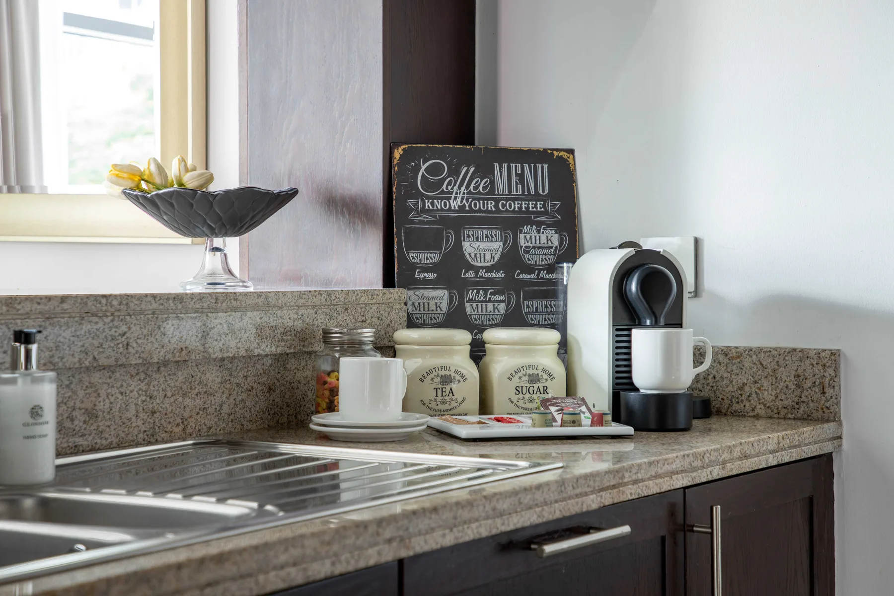 Una encimera de cocina con cafetera, tazas y fregadero.