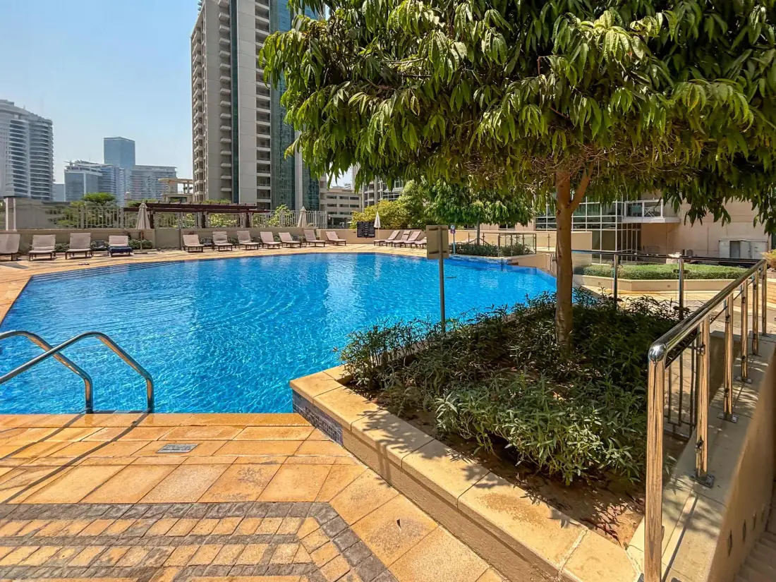 Una piscina junto a una terraza pavimentada, jardineras y edificios altos.