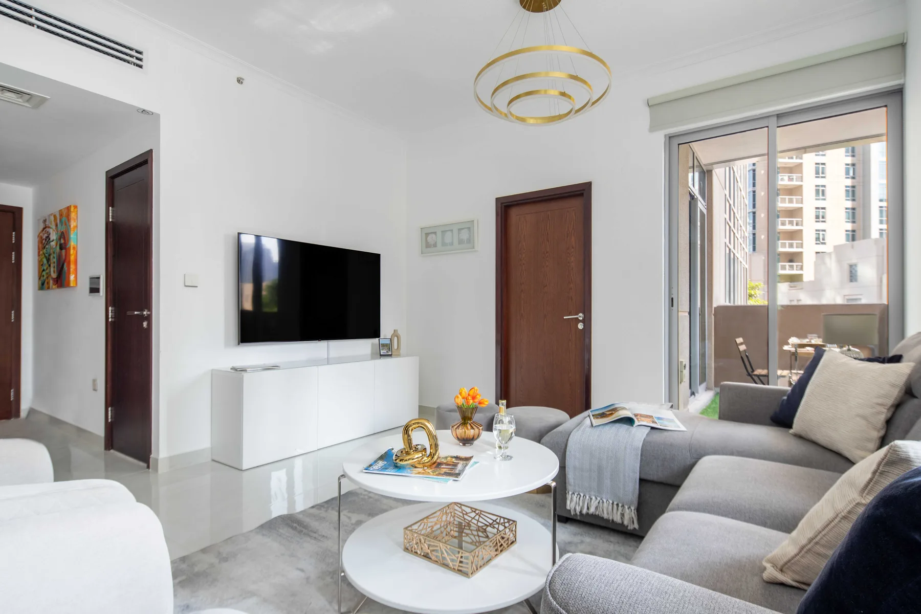 Una sala luminosa con televisor, asientos grises y una puerta de vidrio al balcón.