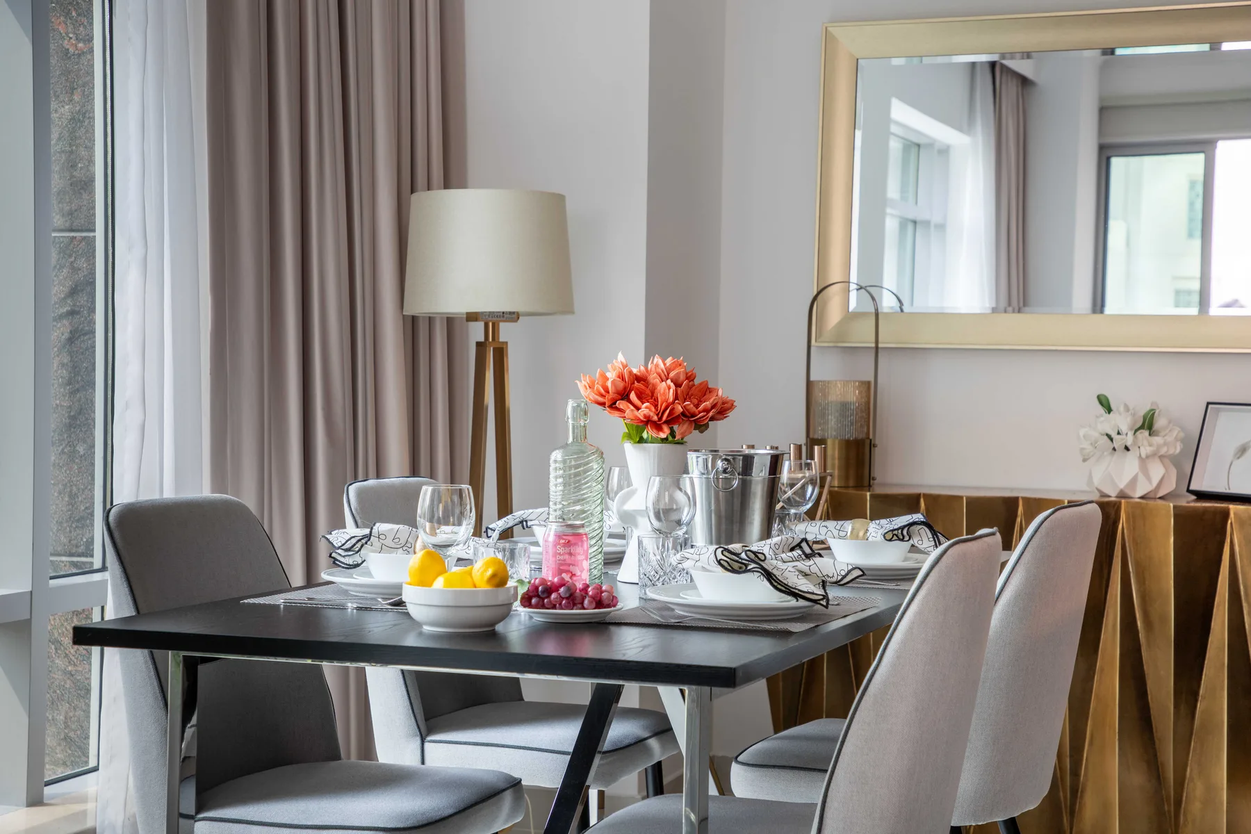 Un comedor con una mesa decorada con flores y fruta junto a ventanas altas.