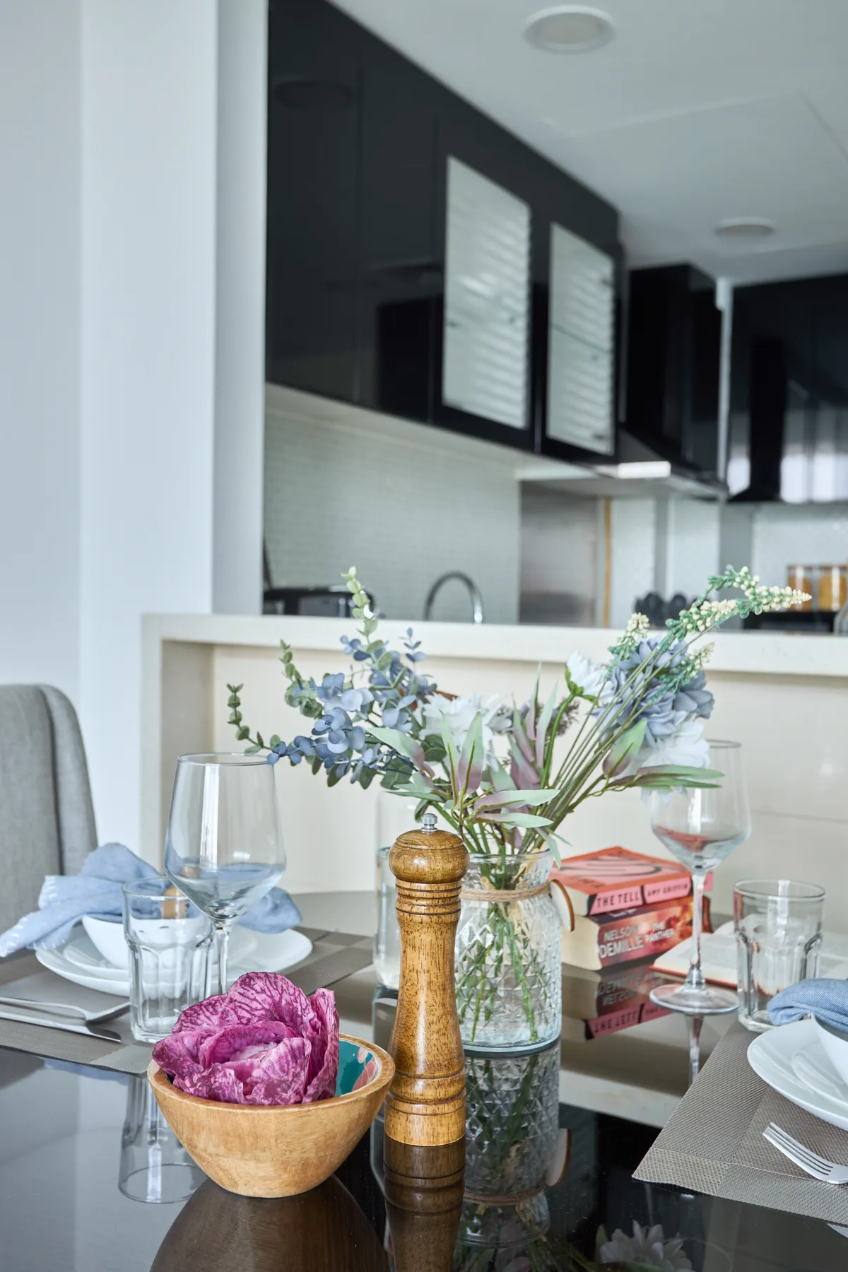Una mesa de comedor con un arreglo floral y cubiertos está frente a una cocina moderna.