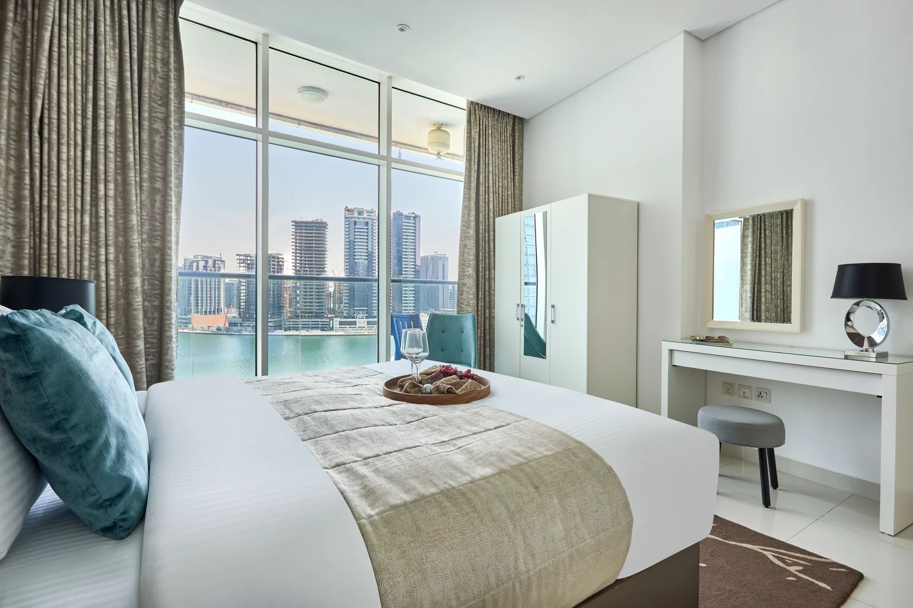 Un dormitorio luminoso con una cama bien tendida junto a grandes ventanales con vistas a la ciudad.