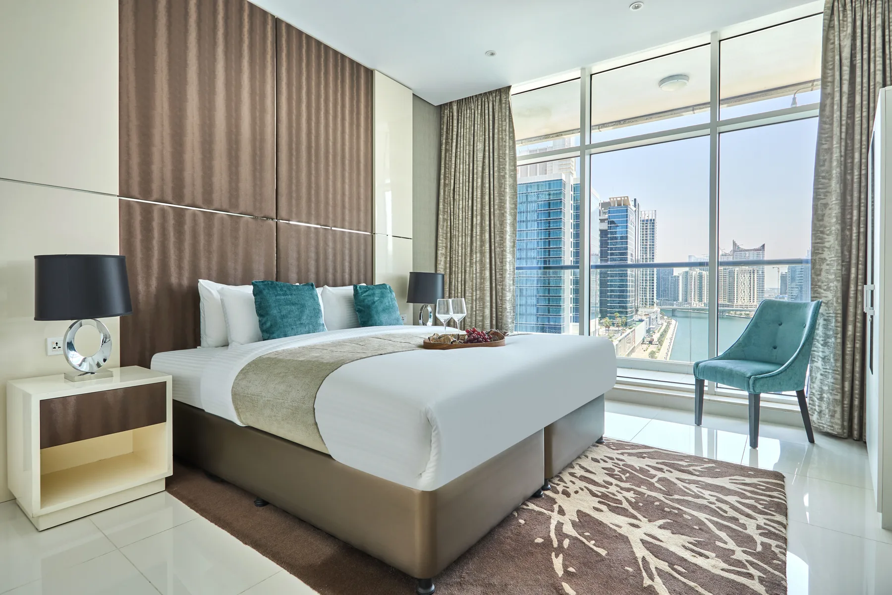 Un dormitorio luminoso con una cama bien tendida junto a ventanales con vistas a edificios de la ciudad.