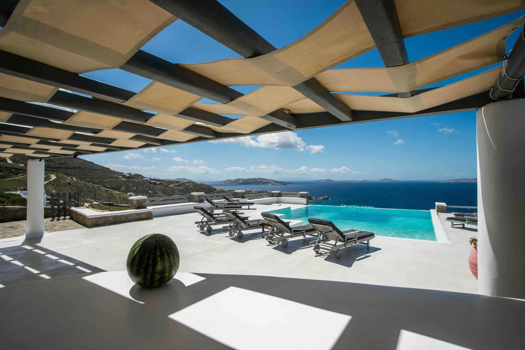 An outdoor pool terrace overlooks the sea, with lounge seating beneath a shaded pergola.