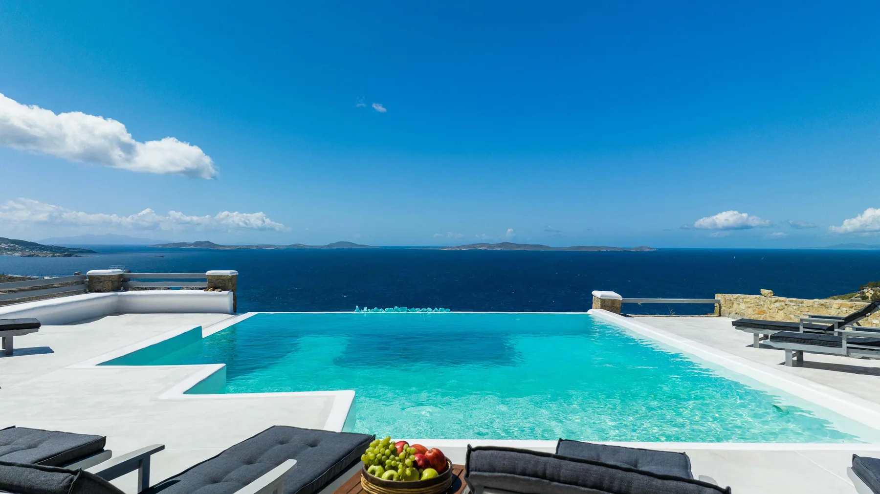 An outdoor swimming pool overlooks the sea beneath a clear blue sky.