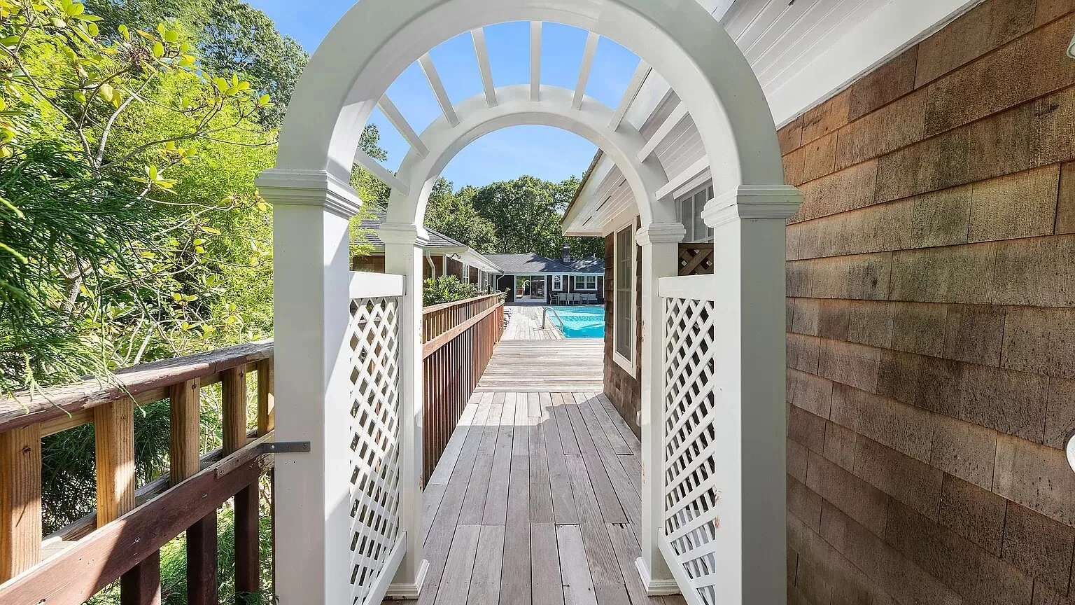 A wooden deck walkway passes through a white arched trellis beside a swimming pool.