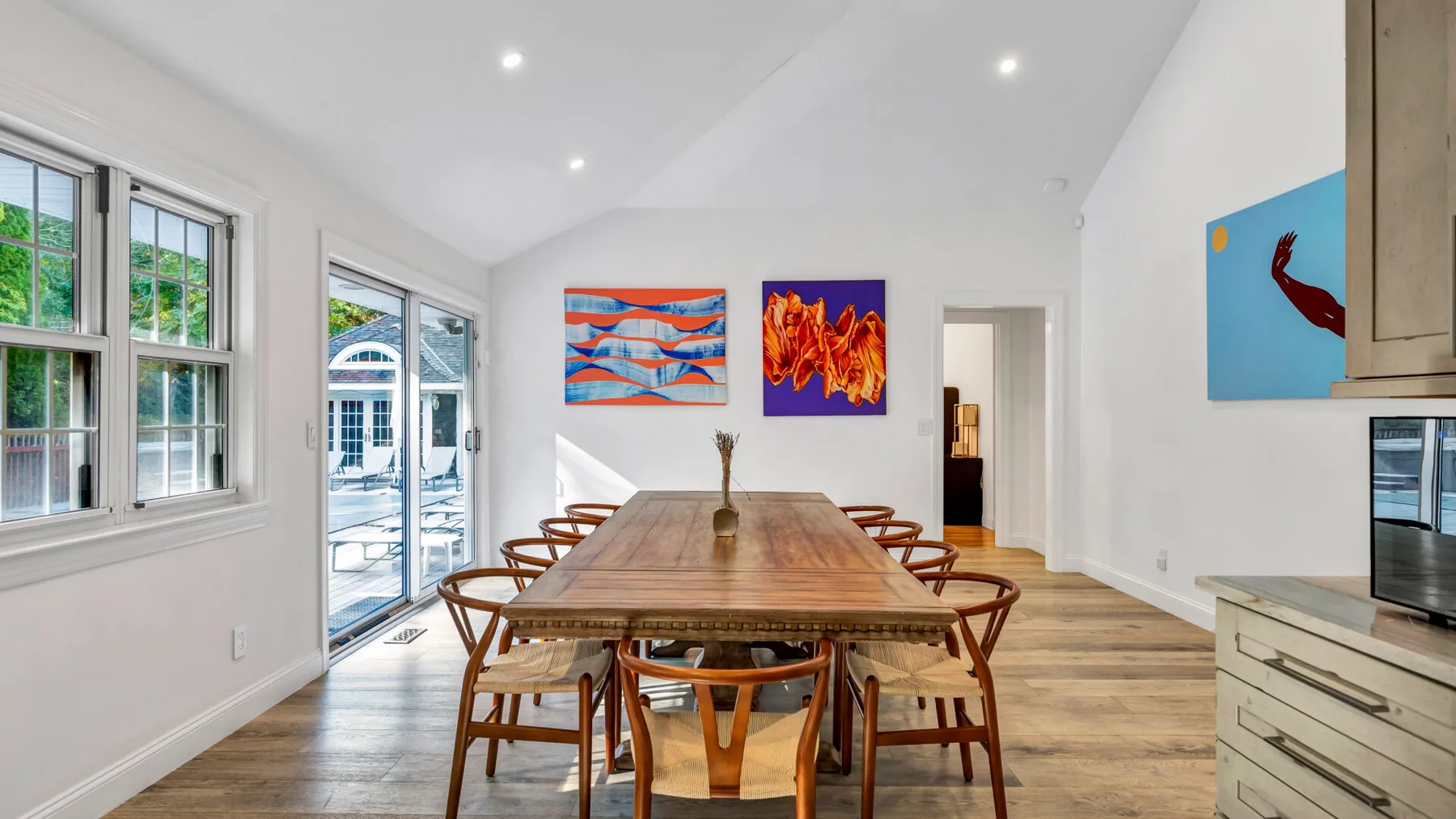 A bright dining room with a long wooden table beneath colorful wall art.