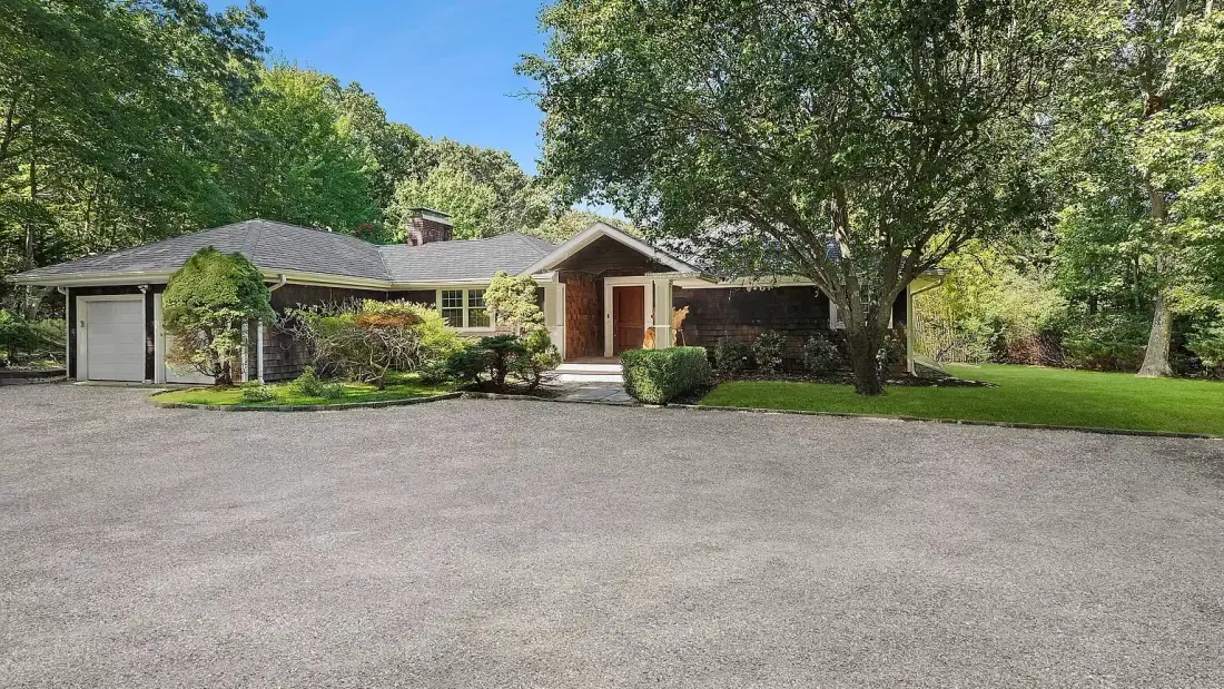 A single-story house sits beside a broad paved driveway and mature trees.