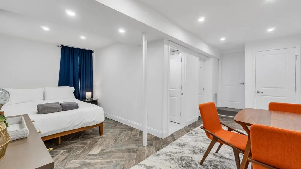 A bright bedroom and dining area with a bed, orange seating, and several doors.