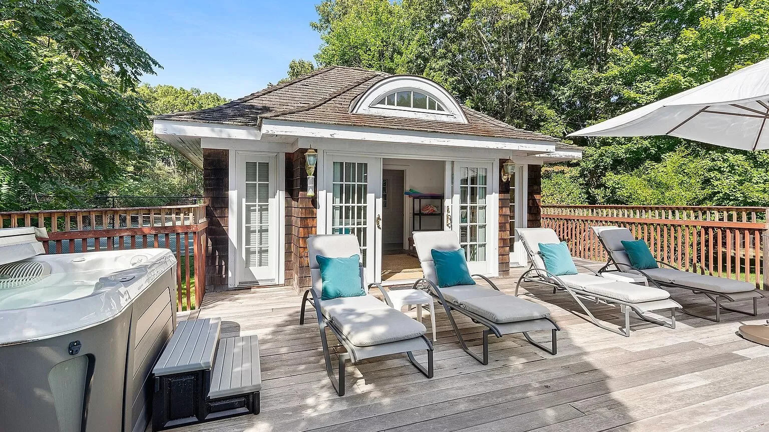 A sunny outdoor deck with lounge seating, a hot tub, and doors opening to a small pavilion.