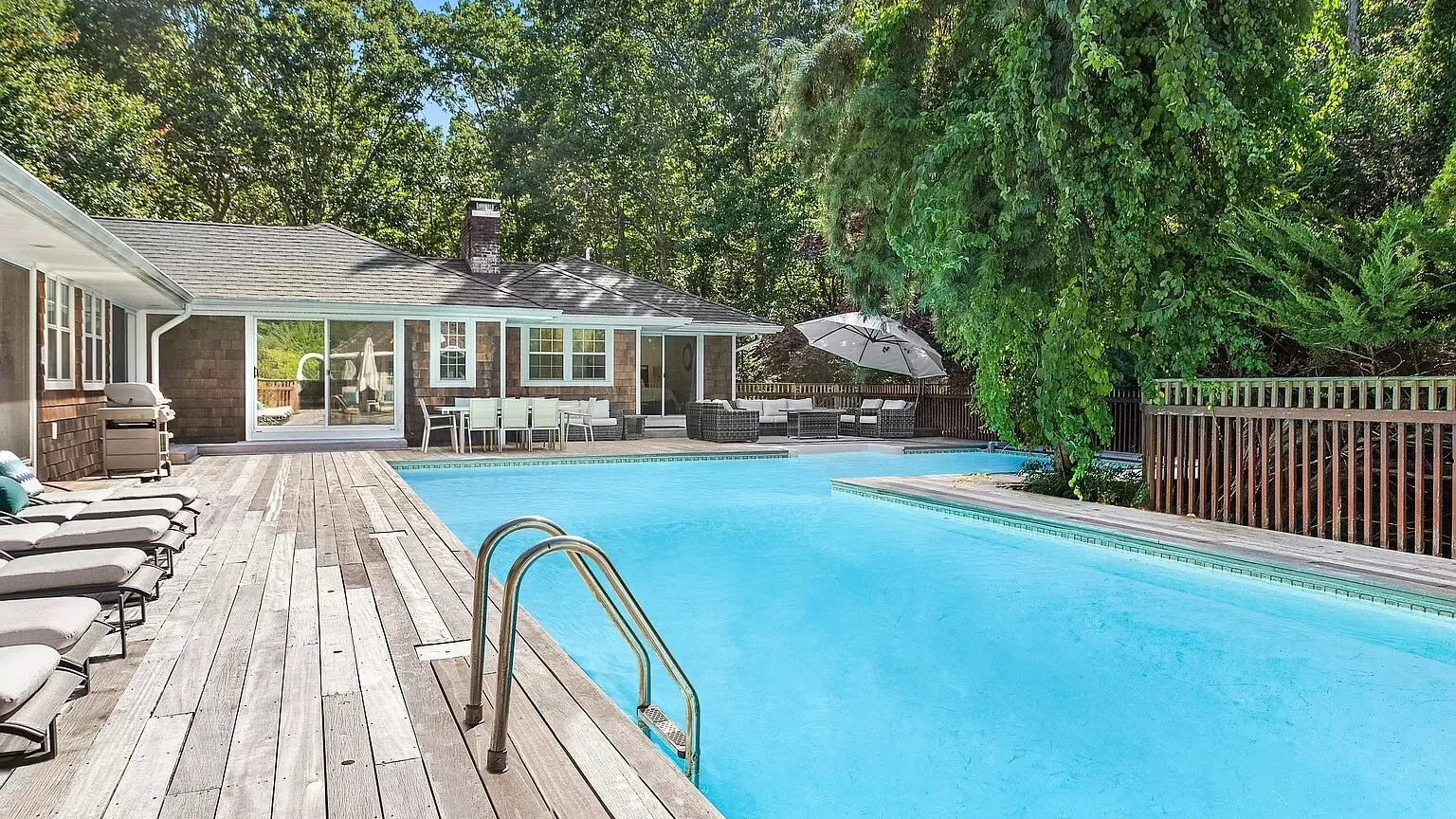 A backyard swimming pool sits beside a wooden deck and a low house.