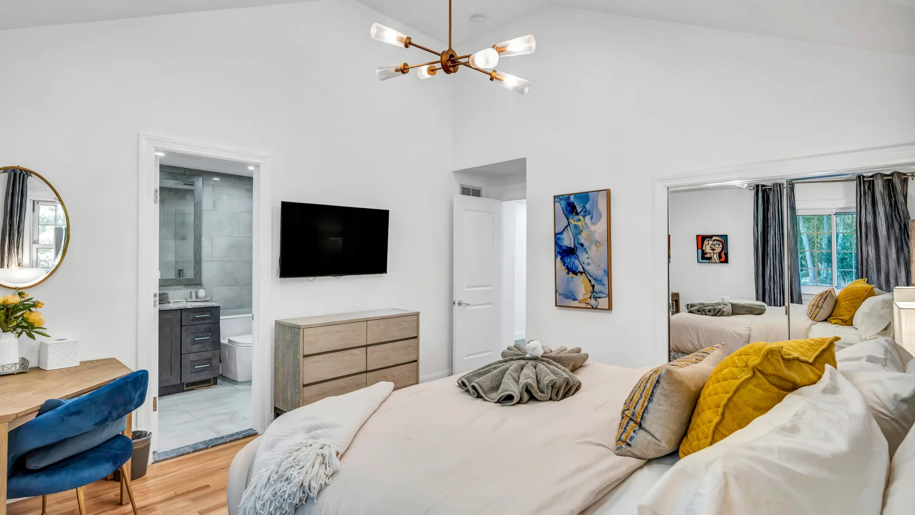A bright bedroom with a large bed, a dresser, and a wall-mounted television.