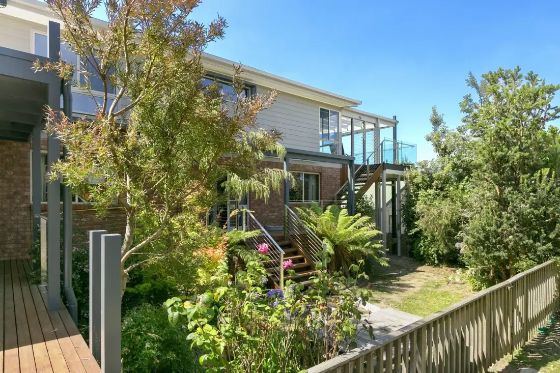 A modern house with an elevated deck, stairs, and a leafy garden.