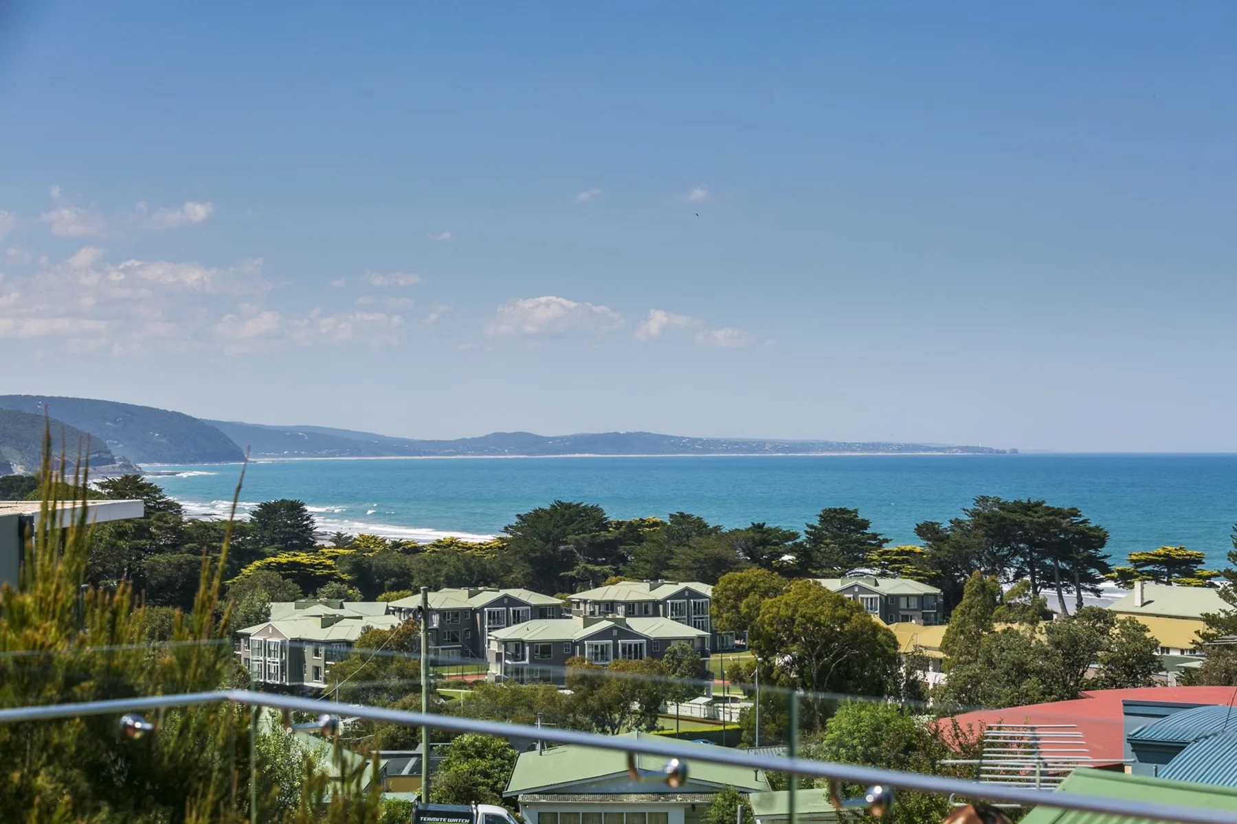 Coastal view of beachfront homes, trees, and the ocean beneath a clear sky.