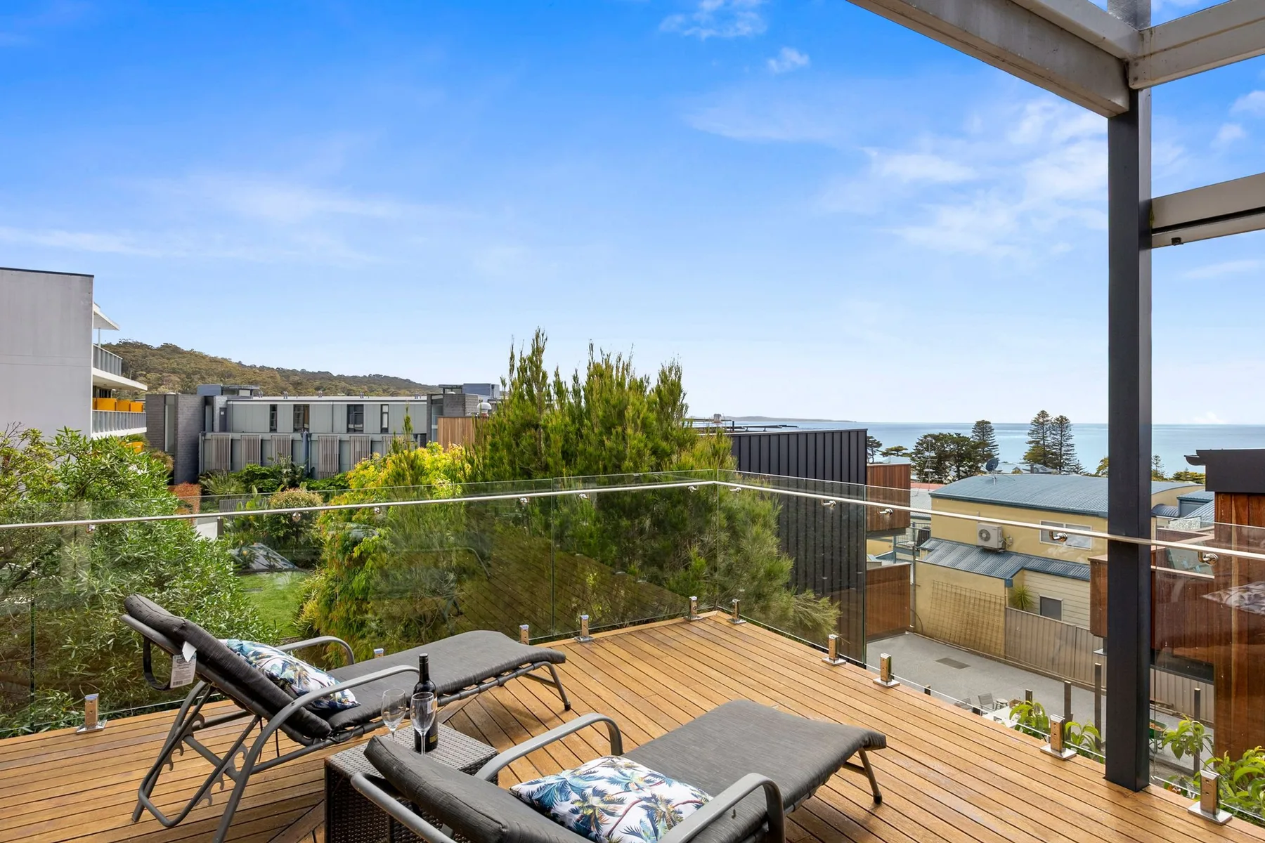 A wooden deck with outdoor seating overlooks nearby buildings and the ocean.