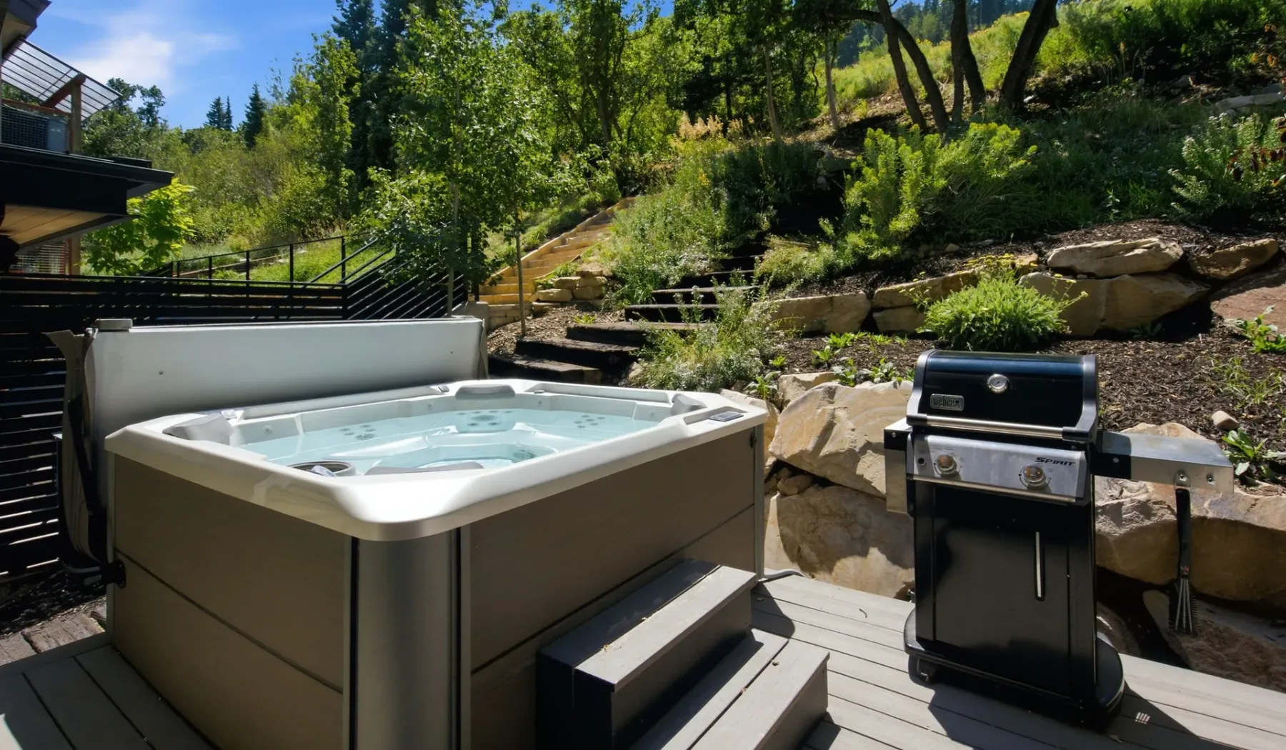 Una bañera de hidromasaje y una parrilla están en una terraza junto a una ladera ajardinada.