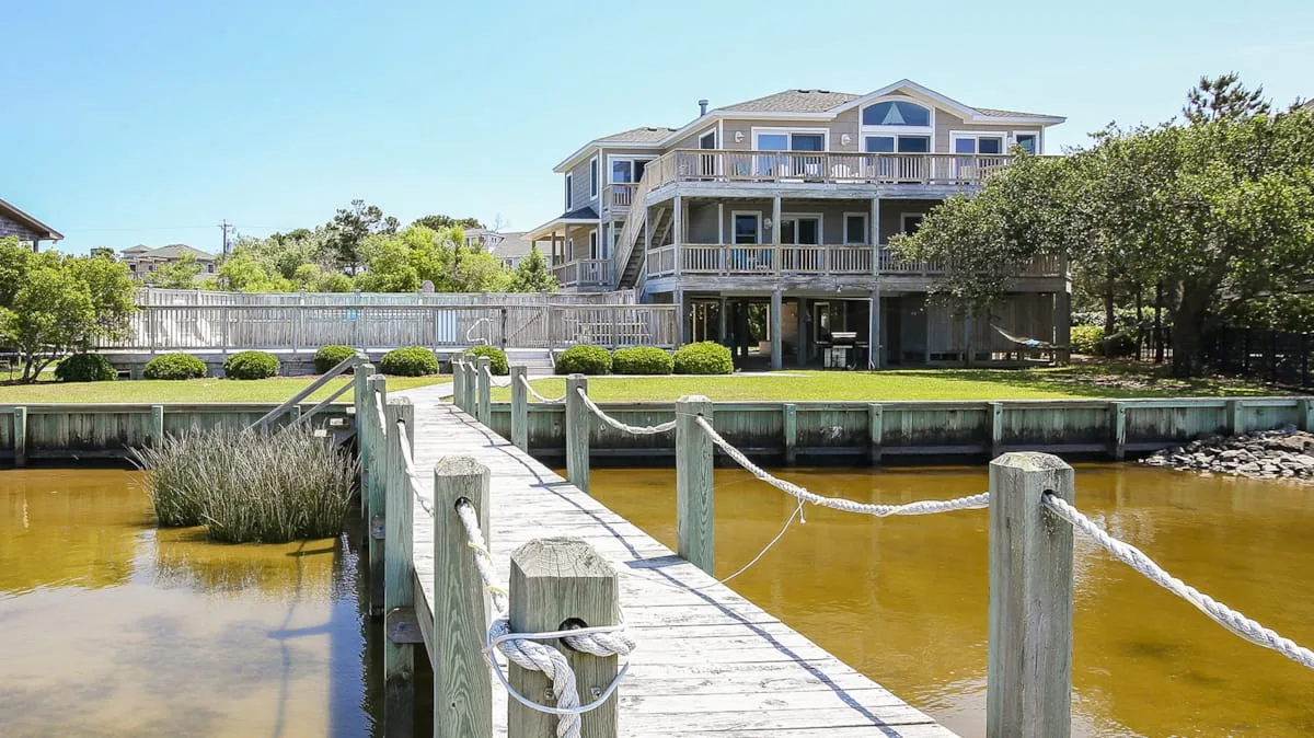 A large waterfront house stands beyond a wooden dock over a shallow waterway.