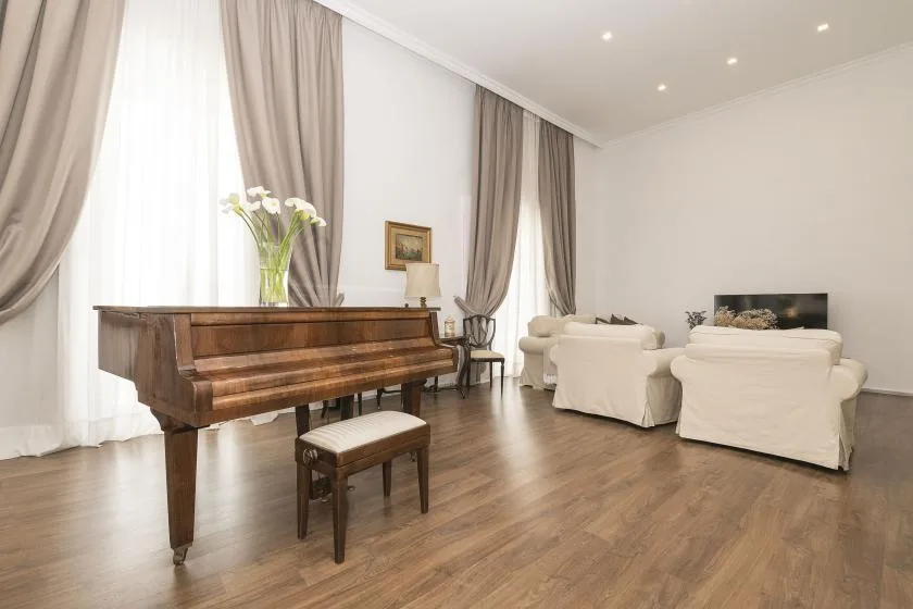 A bright living room with a grand piano and seating beside tall curtained windows.