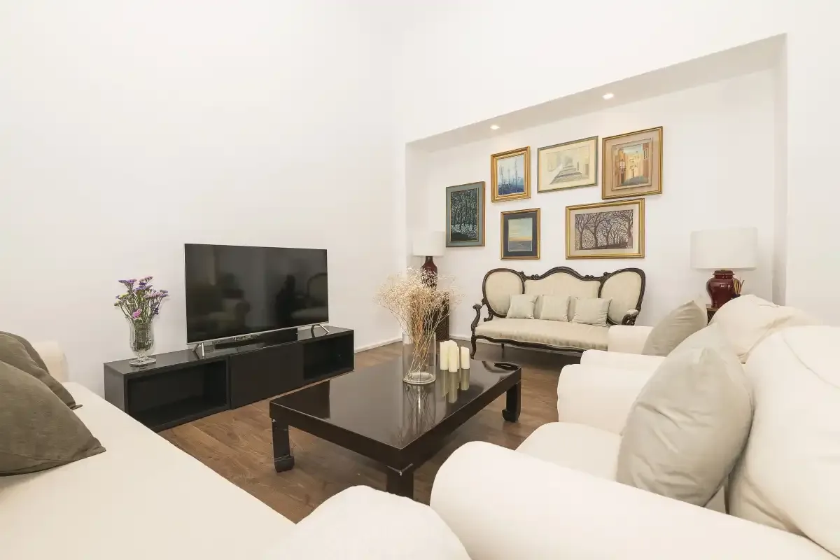 A bright living room with light-colored seating, a television, and framed pictures.