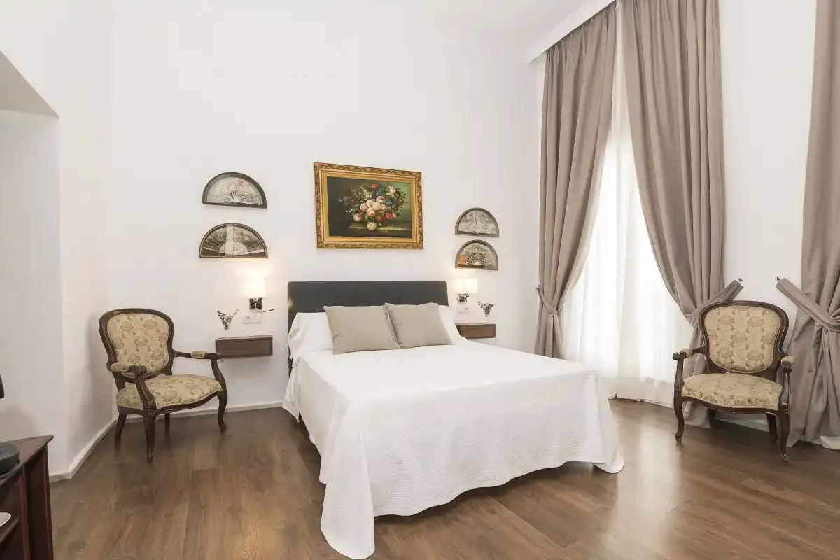 A bedroom with a neatly made bed, two upholstered chairs, and tall curtains.