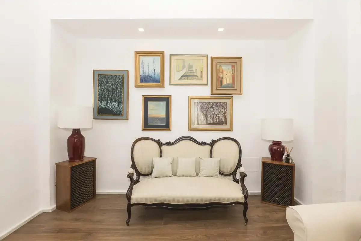 A bright sitting room with an ornate settee beneath a gallery of framed pictures.