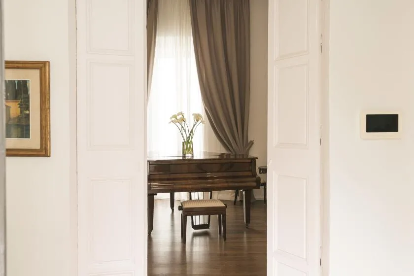 A piano and small stool sit in a bright room viewed through open doors.