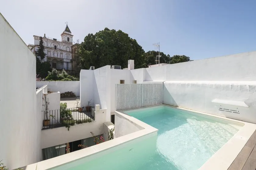 A rooftop pool sits among white walls, with trees and a building beyond.