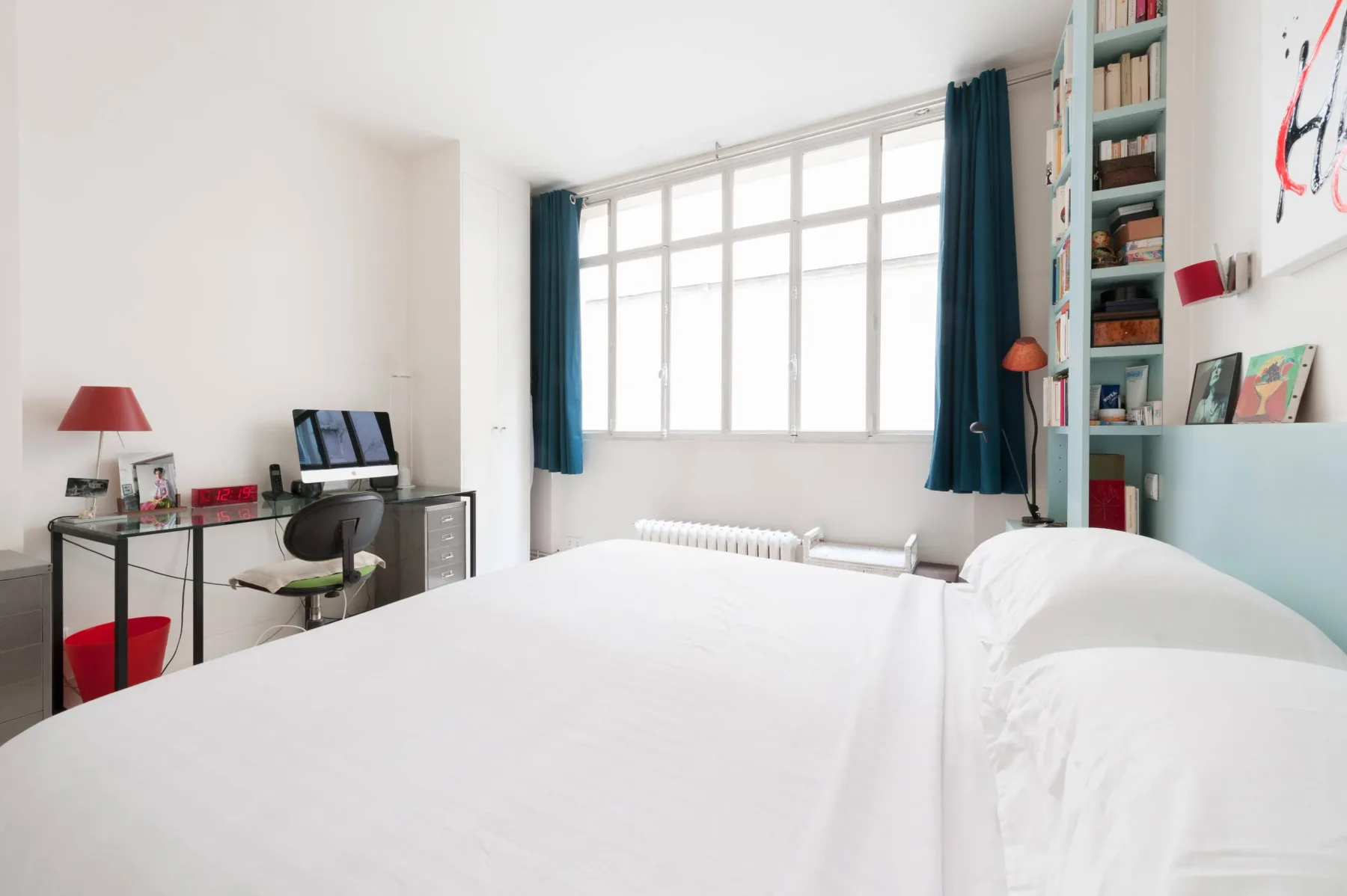 A bright bedroom with a large window, dark curtains, and a white bed.
