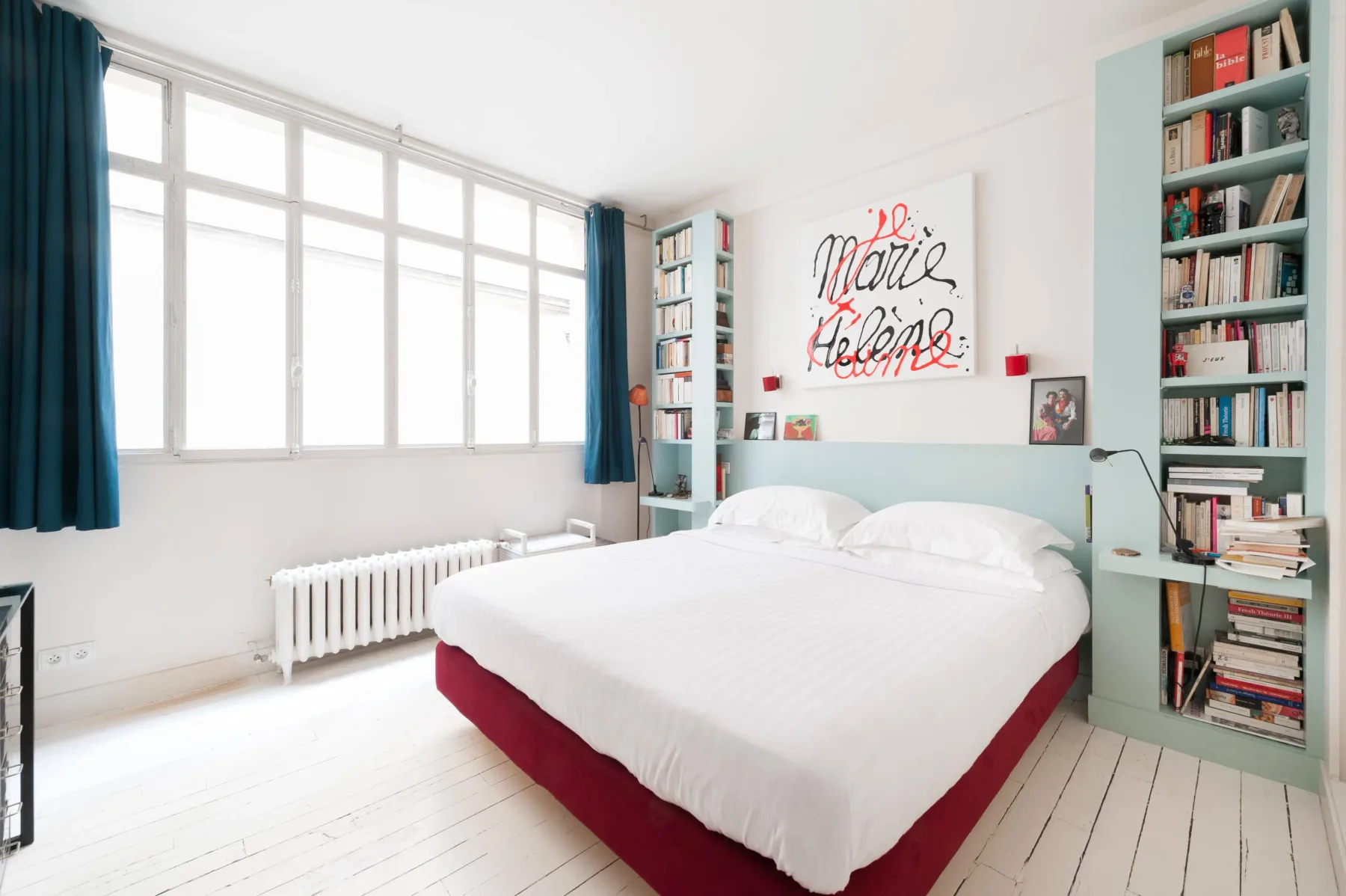 A bright bedroom with a white bed beneath large windows and built-in bookshelves.