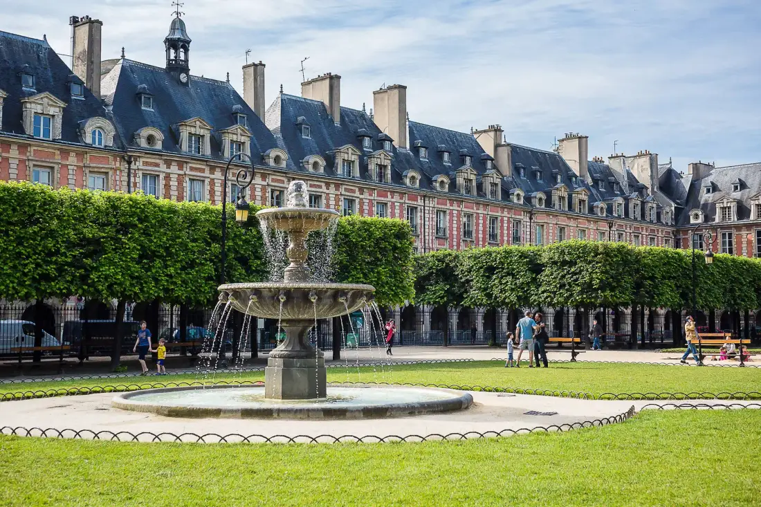 A courtyard fountain stands before a row of historic buildings and trimmed trees.