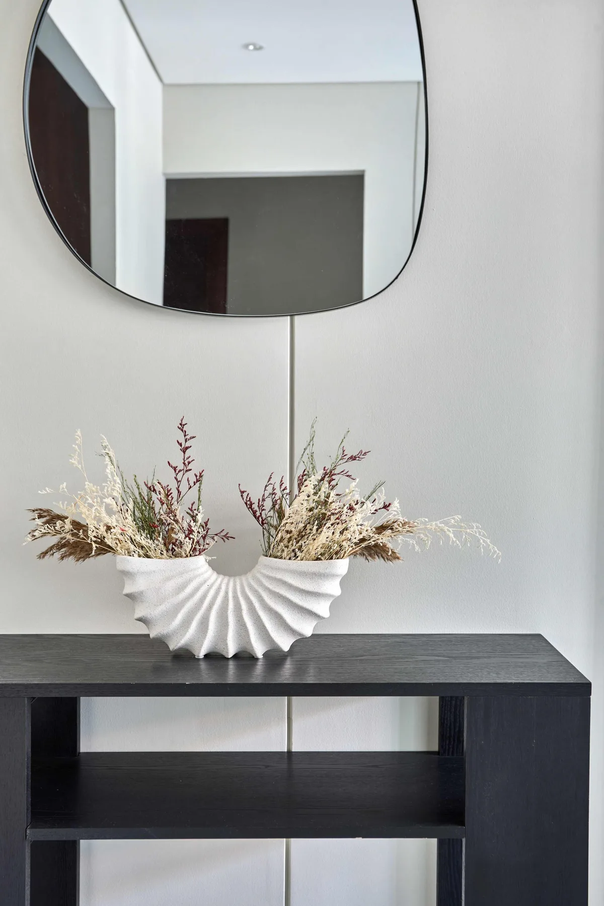 A decorative arrangement of dried flowers sits on a dark console beneath a wall mirror.