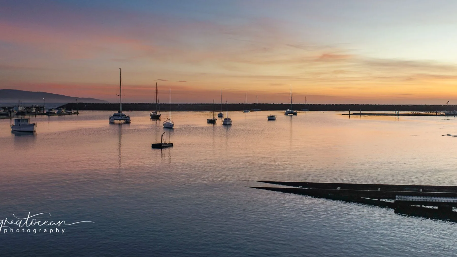 Sailboats sit on calm water beneath a colorful sunset sky.