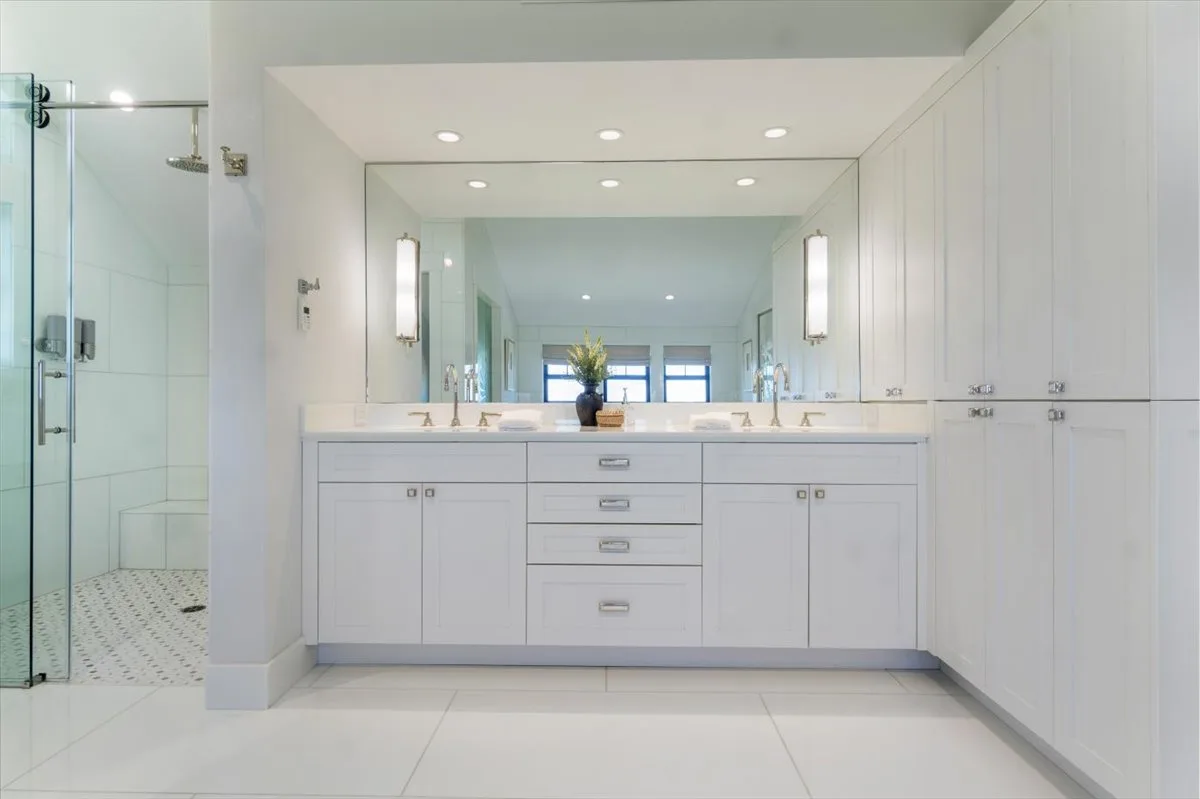 A bright bathroom features a wide vanity beneath a large mirror, with a glass shower beside it.