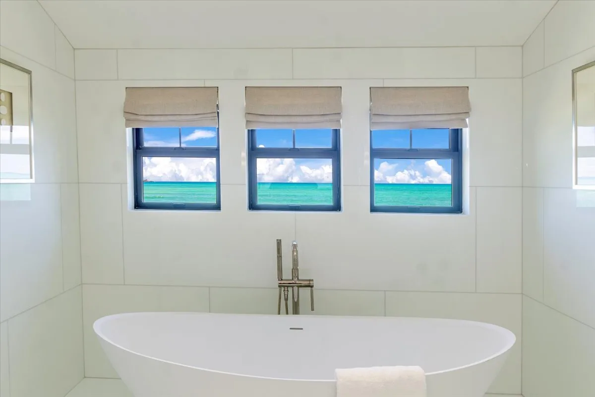 A bathtub sits beneath three windows overlooking the ocean.