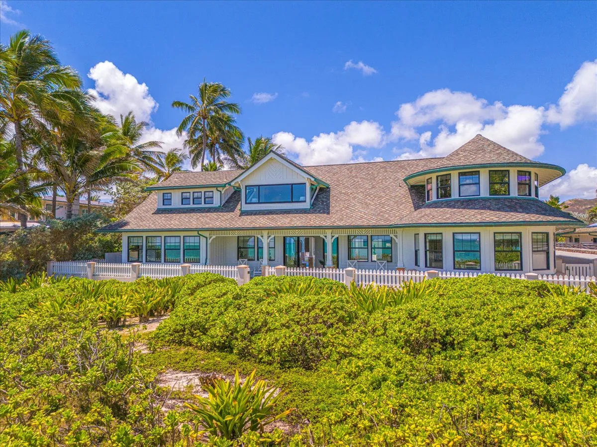 A large house stands behind dense green landscaping and palm trees.