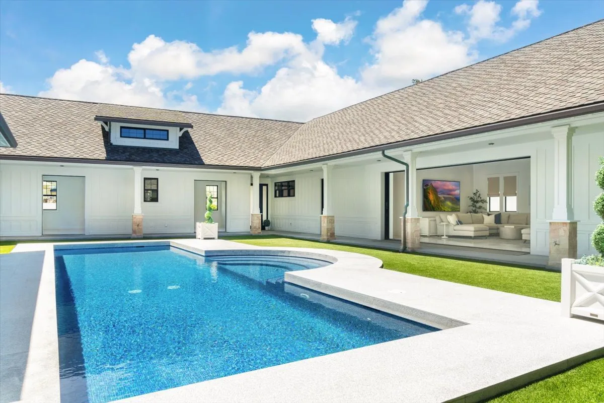 A swimming pool beside a house, with an open living area visible through the windows.