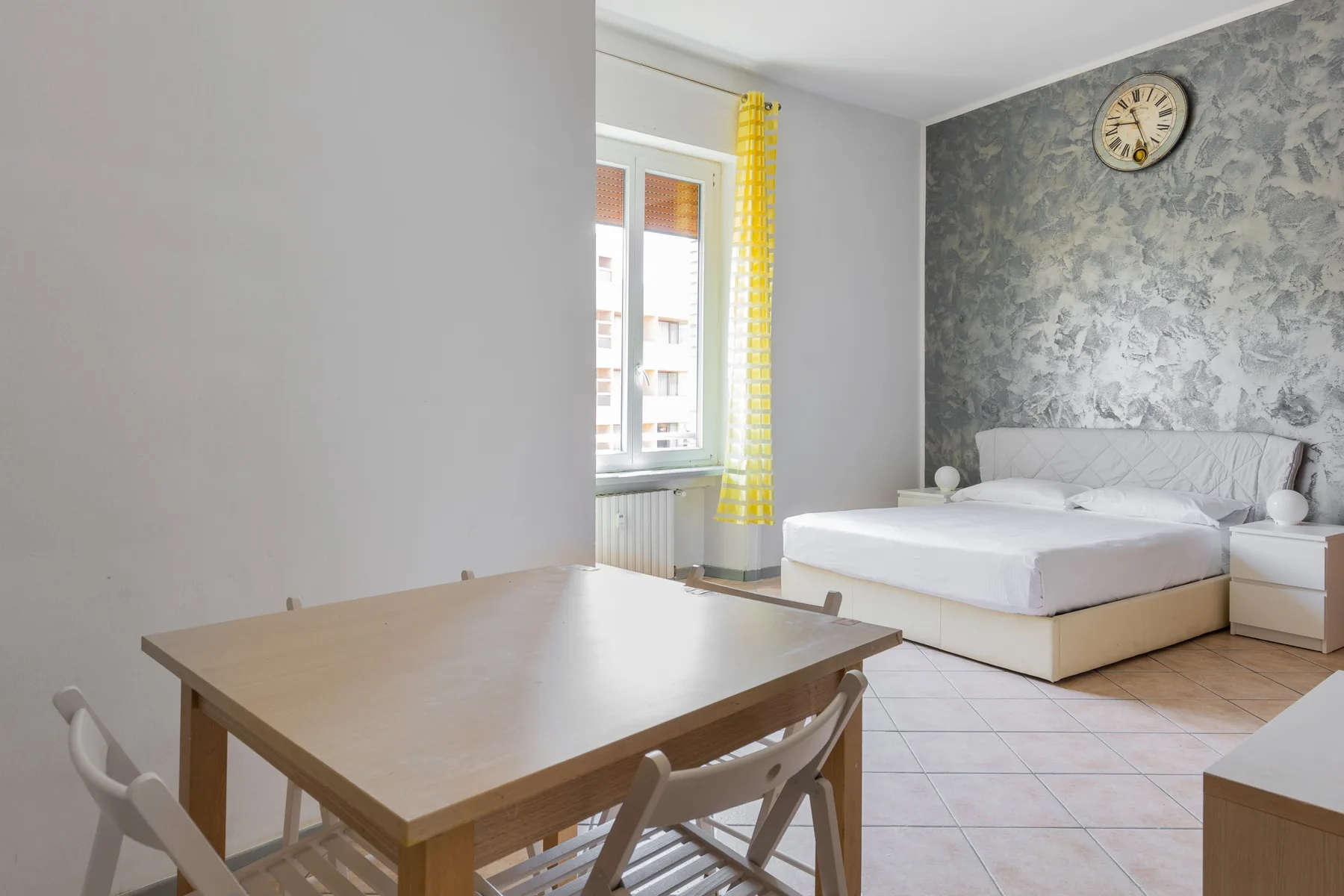 Un dormitorio con una mesa de comedor en primer plano y una cama junto a la ventana.