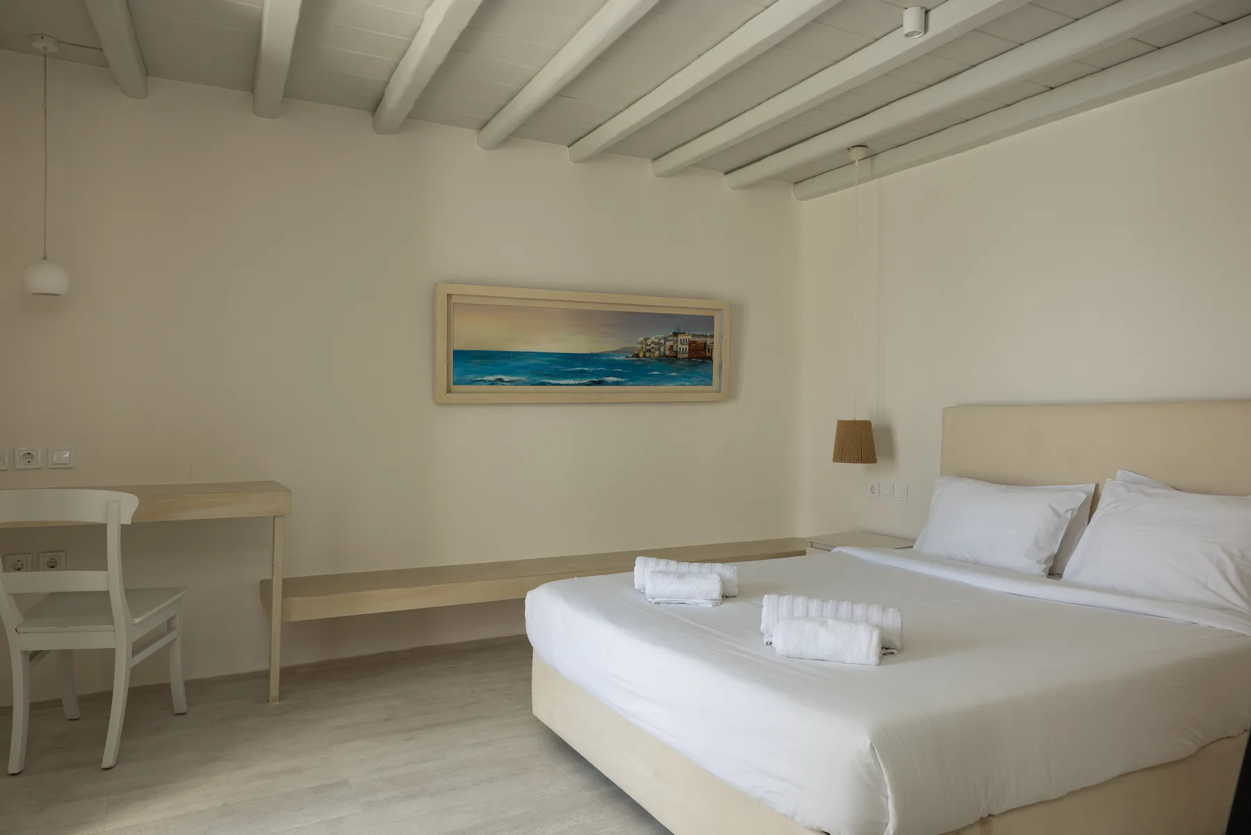 A bright bedroom with white bedding, a desk, and a framed coastal picture.