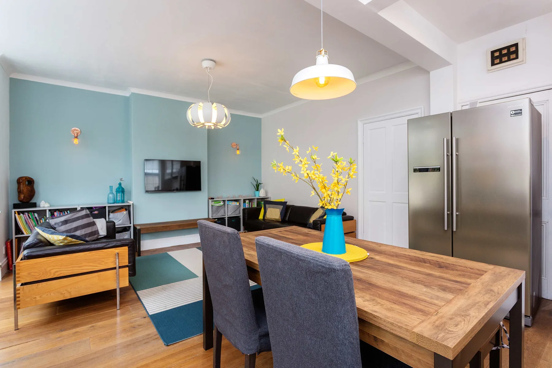 A bright dining and living area with a wooden table, seating, and a wall-mounted television.