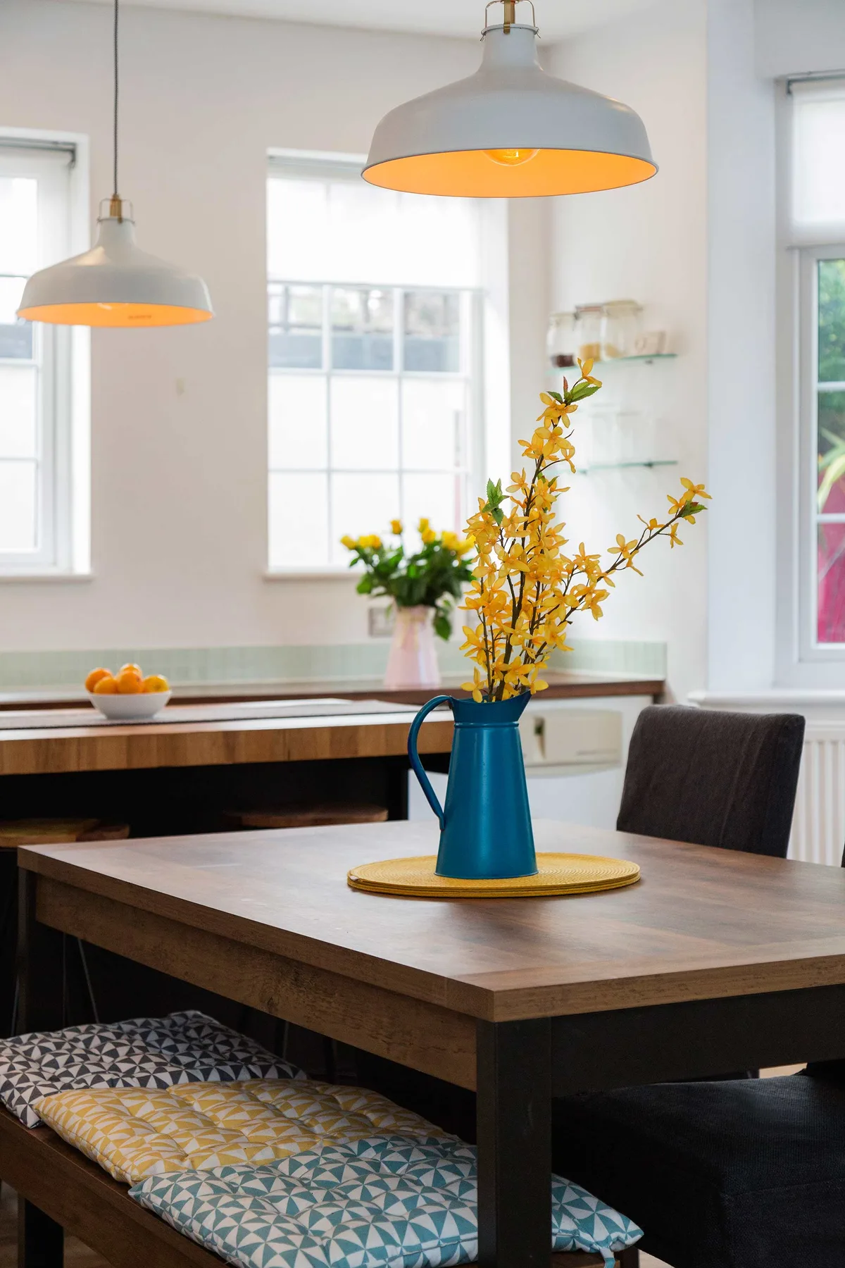 Comedor con una mesa de madera, cojines estampados en un banco y un jarrón con flores amarillas.