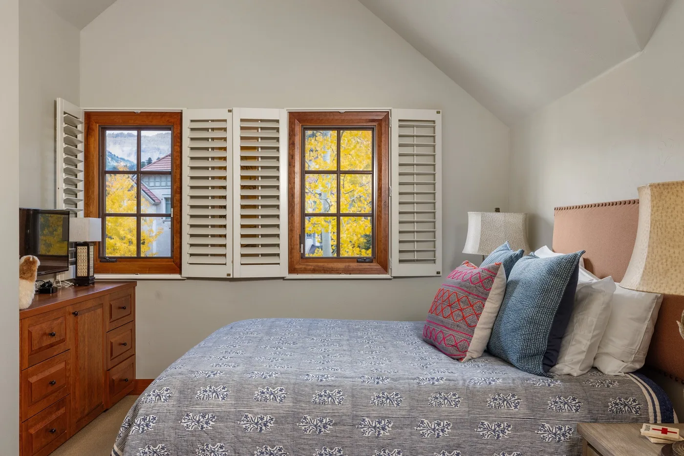 A bedroom with a bed beneath two windows fitted with shutters.
