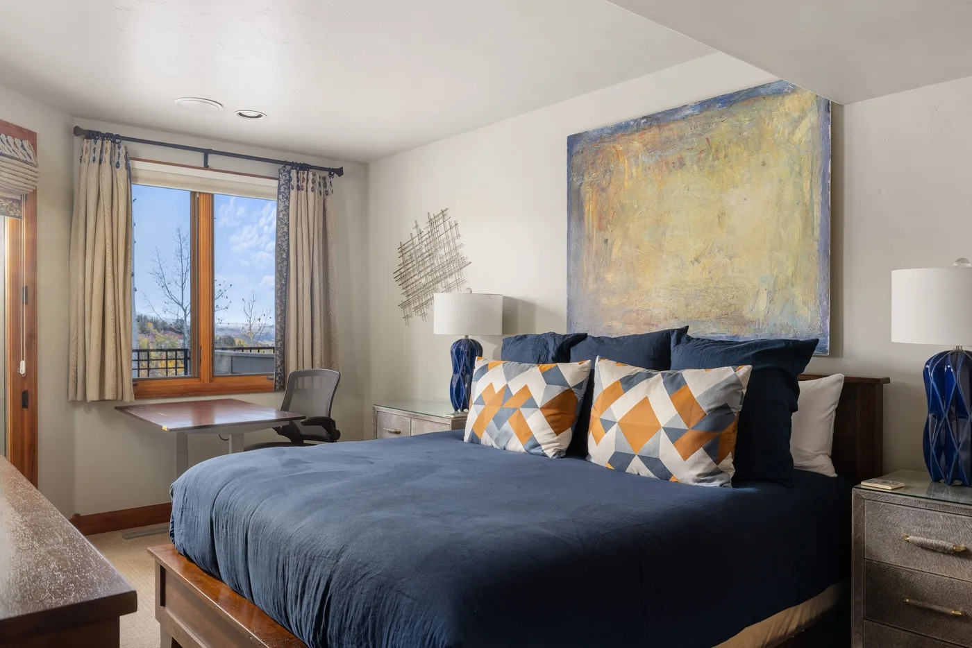 A bedroom with a large bed, patterned pillows, and abstract artwork above the headboard.