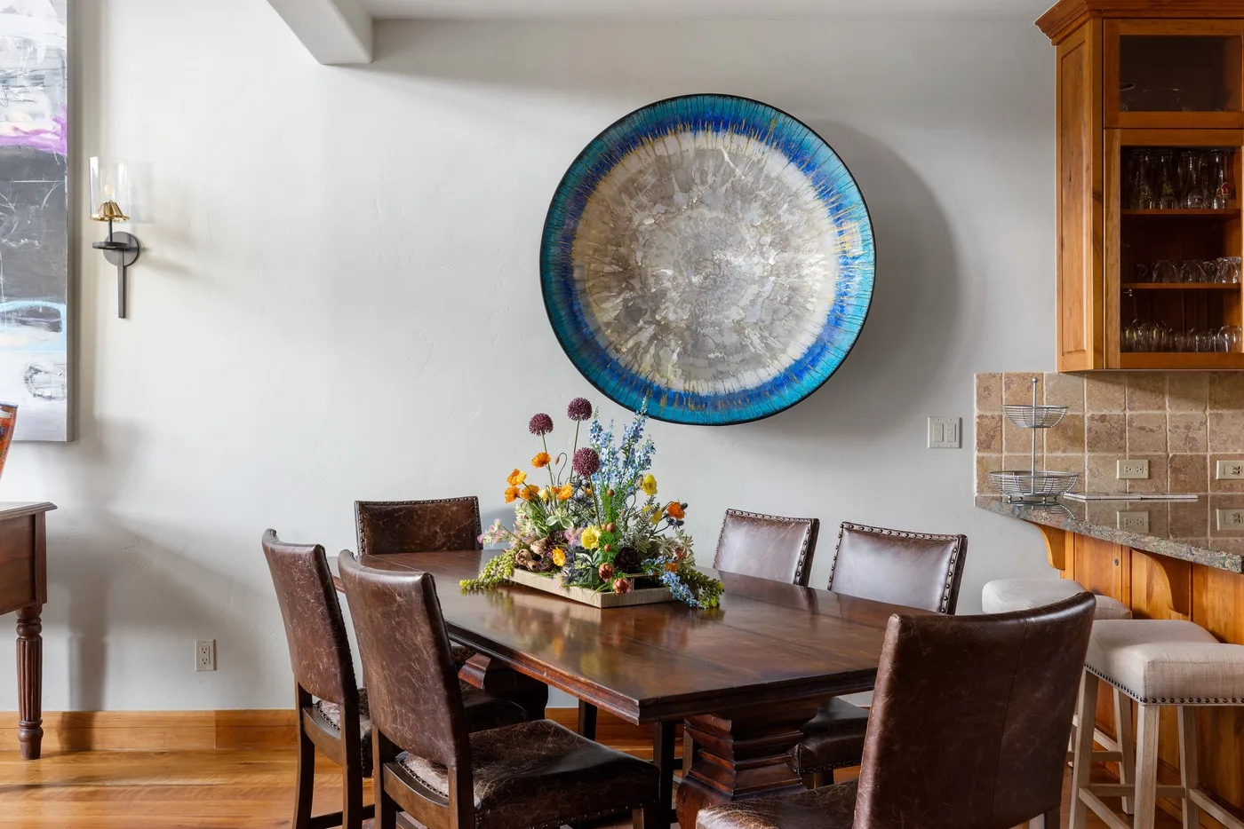 A dining area with a wooden table, chairs, and colorful flowers beneath a circular wall decoration.