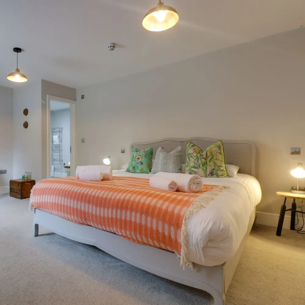 Bedroom with a cane headboard, coral geometric throw and ensuite visible through the open door.