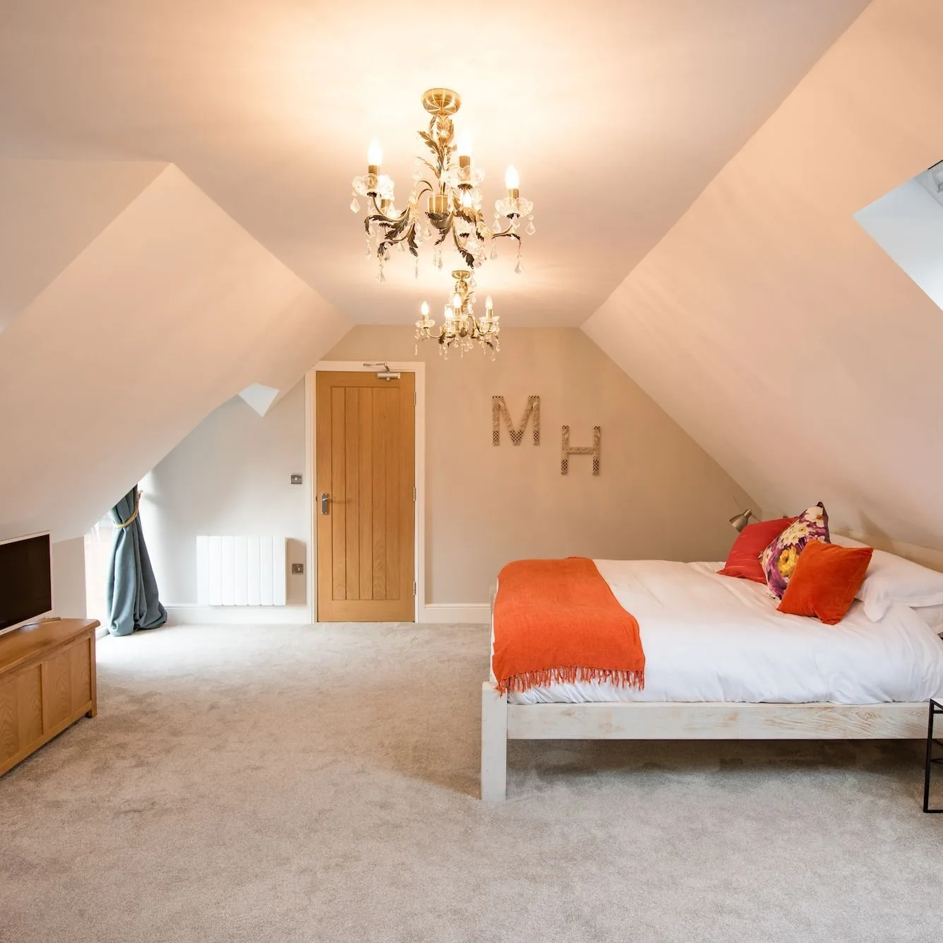 Dormitorio bajo un techo de dos vertientes, con una lámpara de araña de cristal, una manta naranja sobre la cama y letras decorativas de metal en la pared.