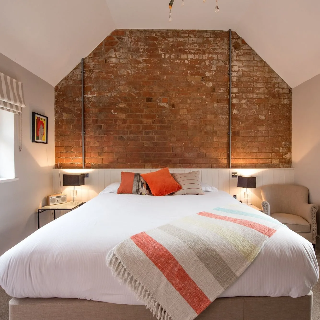 Bedroom with an exposed-brick gable behind the bed, white panelling, orange cushions and a striped throw.