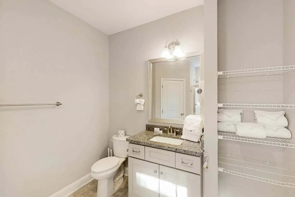 Un tocador de baño con espejo está junto a un inodoro y estantes abiertos con toallas.