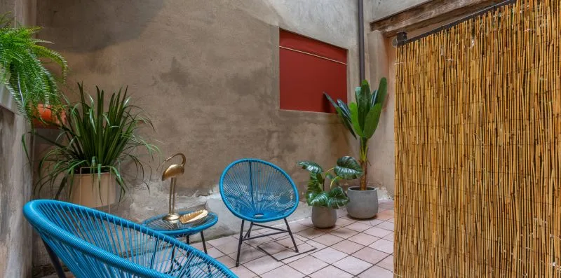 Un patio con suelo de baldosas, asientos azules y plantas en macetas junto a una pantalla de bambú.