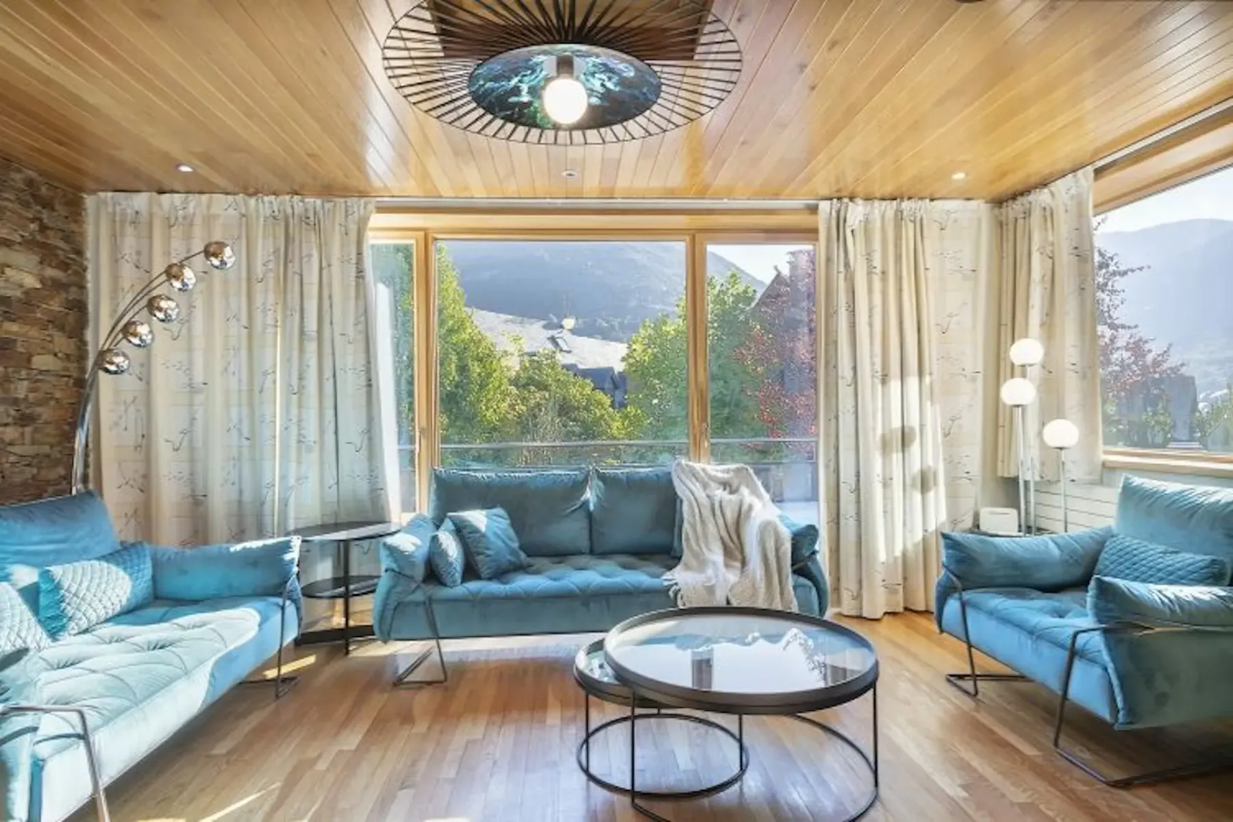 Bright living room with blue sofas, circular coffee tables, and a wooden ceiling. Large windows offer mountain views and natural light.