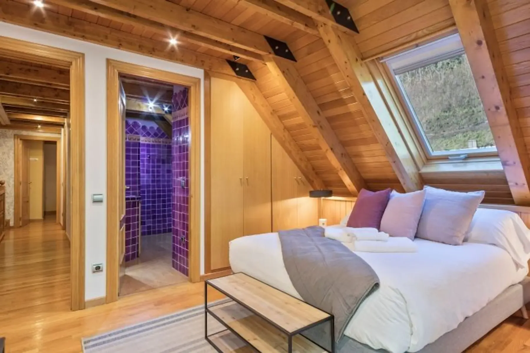 Cozy attic bedroom with wooden beams and sloped ceiling. Features a double bed with pillows, a skylight, and an en-suite bathroom with purple tiles.